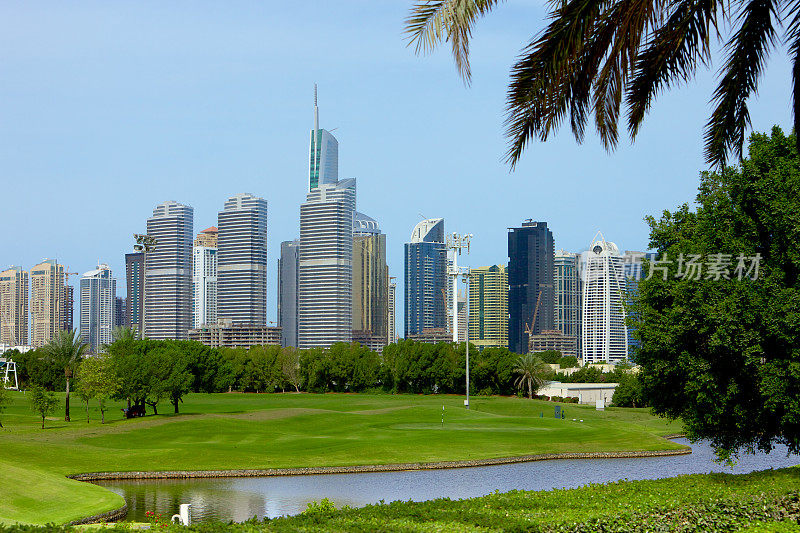 阿拉伯联合酋长国迪拜:高尔夫球道和水的前景;现代摩天楼的JLT在背景中，被一棵椰枣树框在右边-豪华的阿拉伯