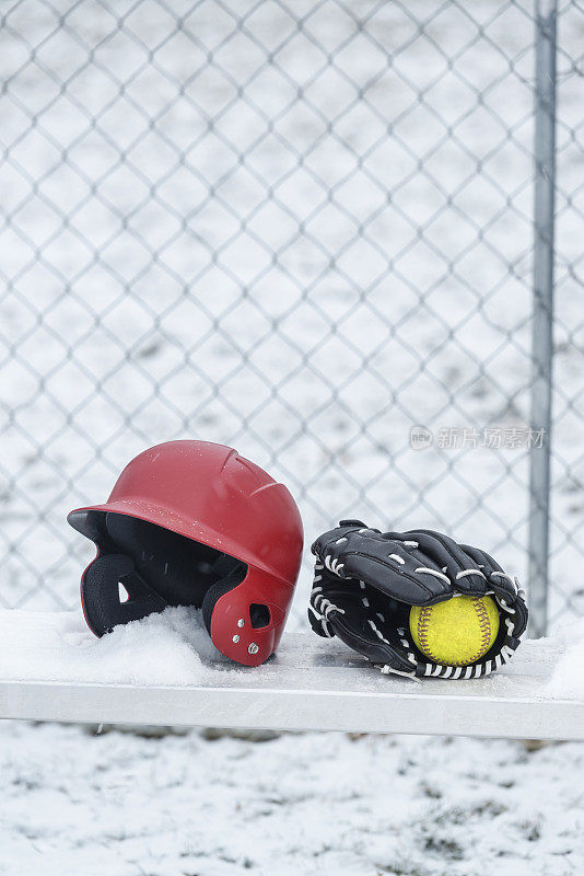 一个戴着垒球手套和棒球头盔的垒球坐在雪地里的长椅上