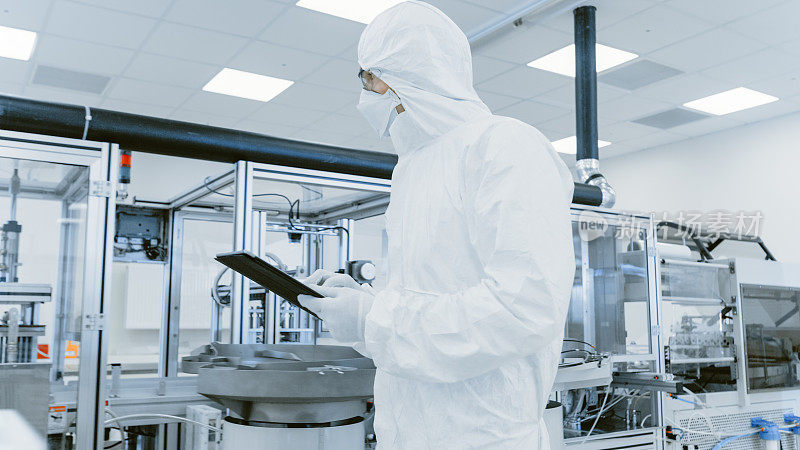 质量控制检查:科学家使用数字平板电脑和穿着防护服走过制造实验室。产品制造:制药，半导体，生物技术。