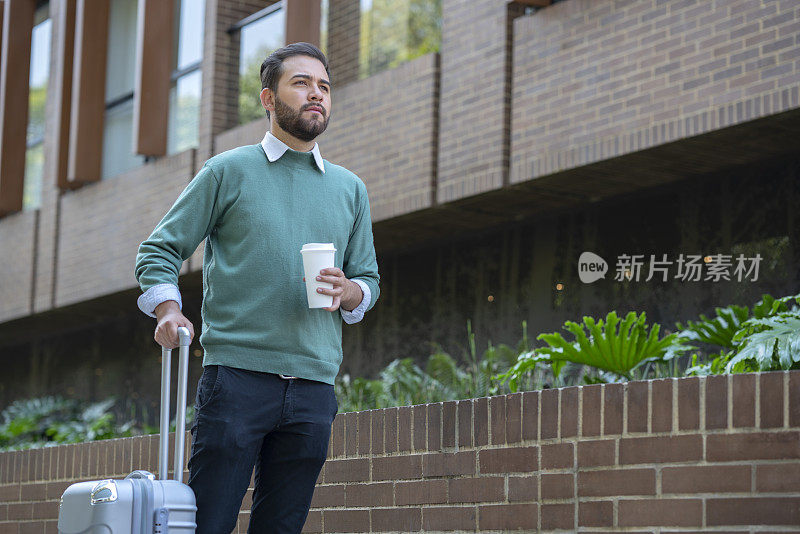 一个拉丁裔的年轻人走在街上，穿着一件绿色毛衣，一只手拿着咖啡，另一只手拿着旅行箱。走出现严重