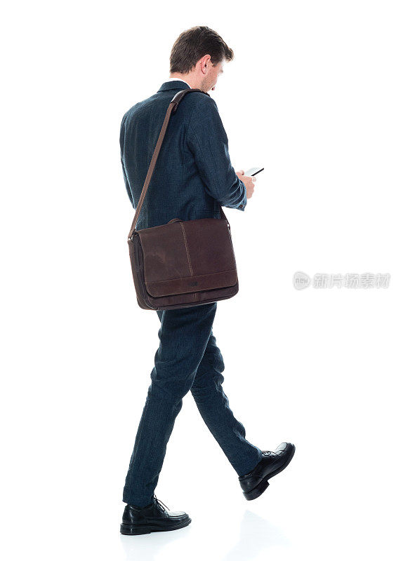 白人男性商务人士在白色背景前走着，背着肩包，拿着包，使用手机