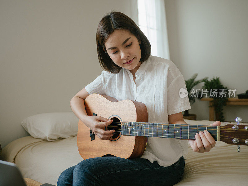 年轻女子微笑着坐在床上拿着吉他看着笔记本电脑