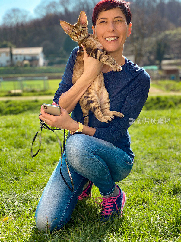 年轻女子抱着大大的微笑和德文郡雷克斯猫在与阳光-股票照片的田野中间摆姿势