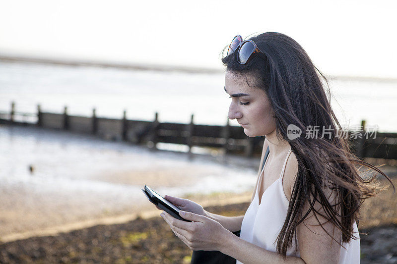 一个十几岁的女孩坐在海滩附近，手里拿着电子阅读器