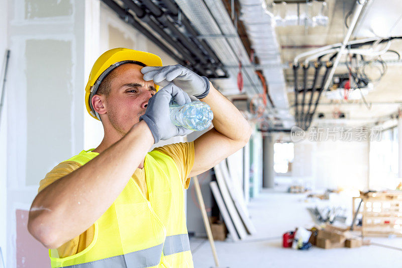 戴着黄色安全帽的年轻建筑工人在建筑工地用一瓶水提神。