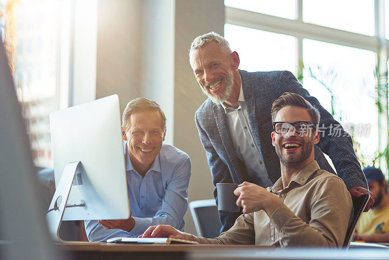 快乐的商务人士。三个开朗的男人坐在办公桌前，用电脑，看着相机，微笑着，在现代化的办公室或联合办公空间里一起做项目