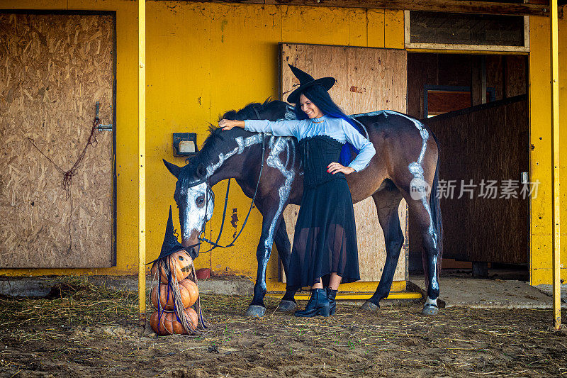 一匹马正在嗅着一个即兴制作的南瓜雕像，一个打扮成女巫的女孩站在旁边微笑着