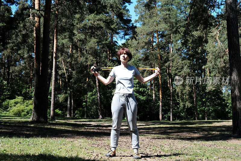 成熟的女人在松林里与阻力带一起锻炼