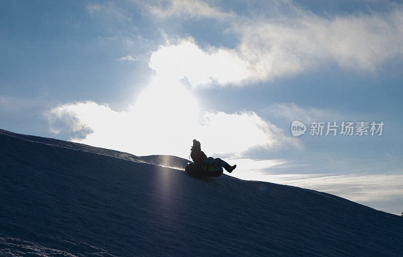 一名年轻女子骑着充气环从雪山上迅速滑下。太阳背景下一个年轻女子的剪影。