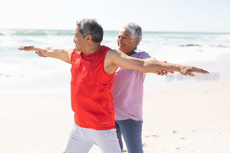 微笑的年长混血女人帮助男人在阳光明媚的海滩上伸展手臂做瑜伽姿势