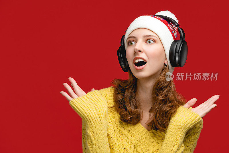 在红色背景的摄影棚里，一幅戴着圣诞老人帽子的善良的成年人肖像正在摆姿势。女人戴着无线耳机，听着新年音乐。与人的概念。