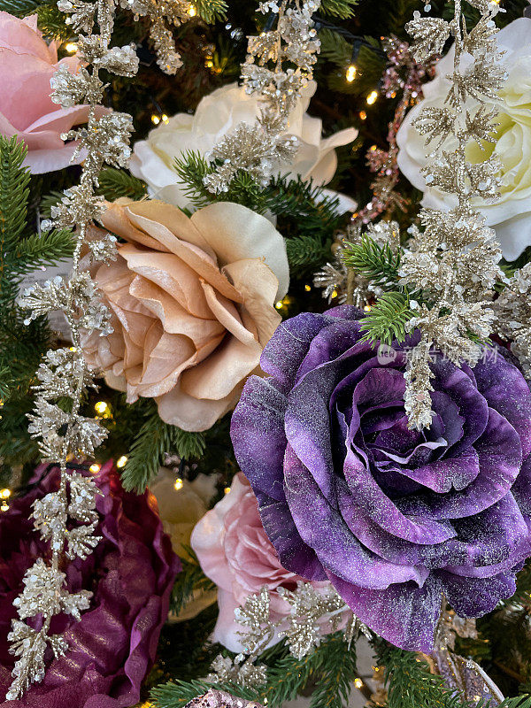 图像以白色、淡粉色、玫瑰金色和紫色玫瑰花冠装饰品挂在人造圣诞树的树枝上，云杉针叶上，模糊的白色神仙灯为背景