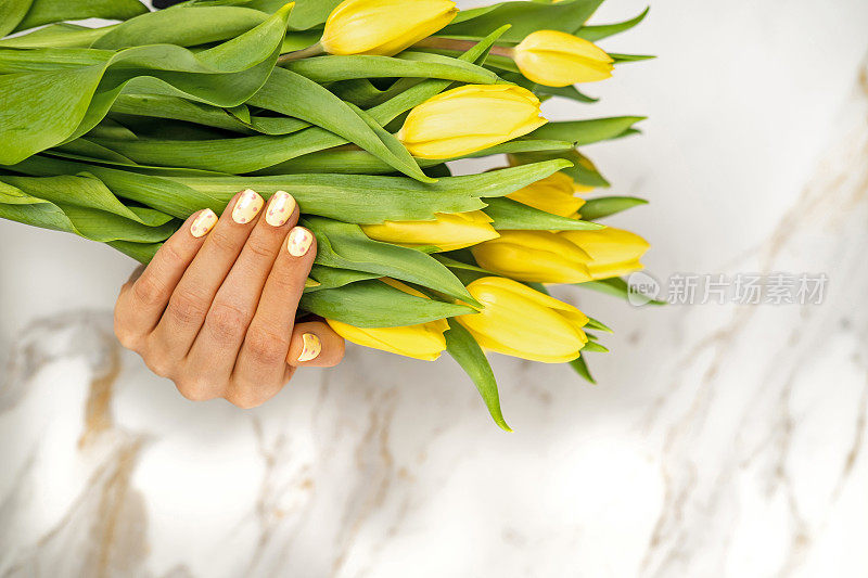 涂着黄色指甲的女人的手拿着白色大理石背景上的一束郁金香。