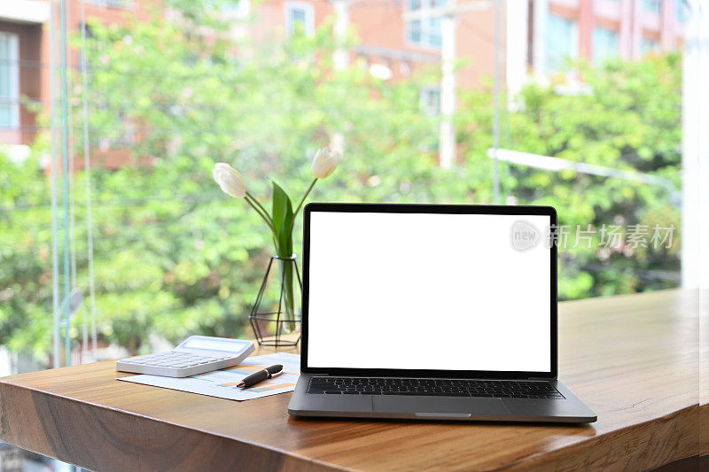 木制桌子上靠近大办公室窗户的白色空屏幕的笔记本电脑模型。