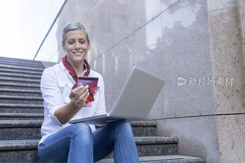 快乐的女人坐在户外的楼梯上，在网上购买时阅读信用卡号码