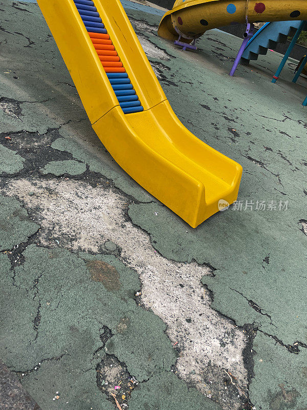 公共公园塑料黄色滑梯游乐设备底部图片，湿淋，橡胶屑和树脂绿色操场地板表面，危险的坑洞和裂缝，关注前景，健康和安全理念
