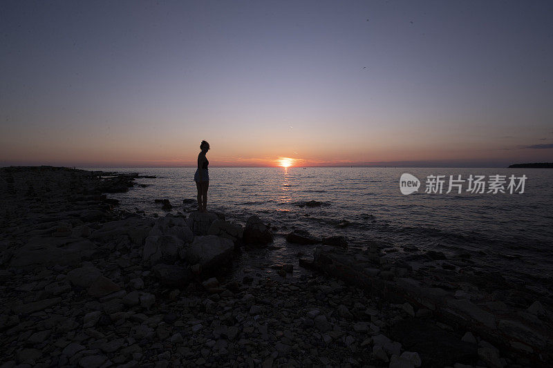 一个女人的剪影，站在海边的一块岩石上，享受着夕阳的余晖