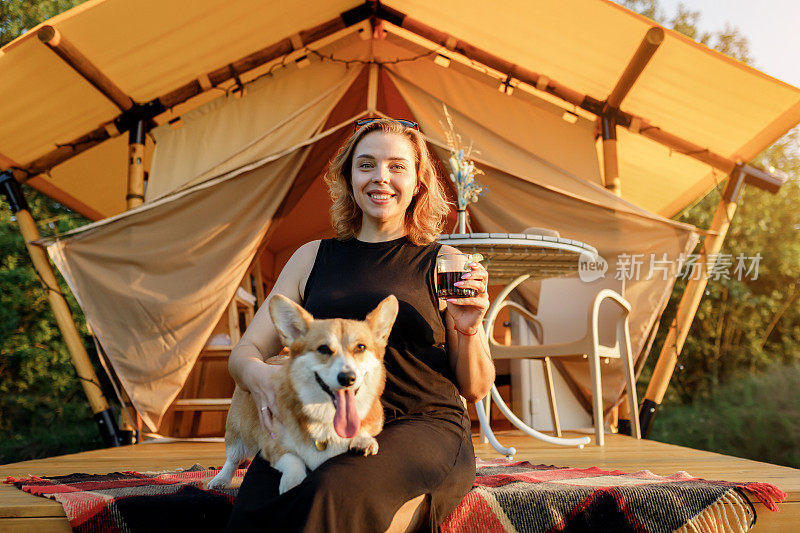 快乐的年轻女子和威尔士柯基彭布罗克犬在夏日的豪华野营中放松。豪华露营帐篷，适合户外休闲娱乐。生活方式的概念