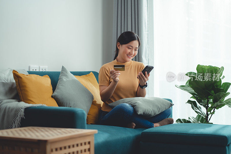 亚洲女性开心兴奋地坐在沙发上用智能手机和信用卡在家网上购物。