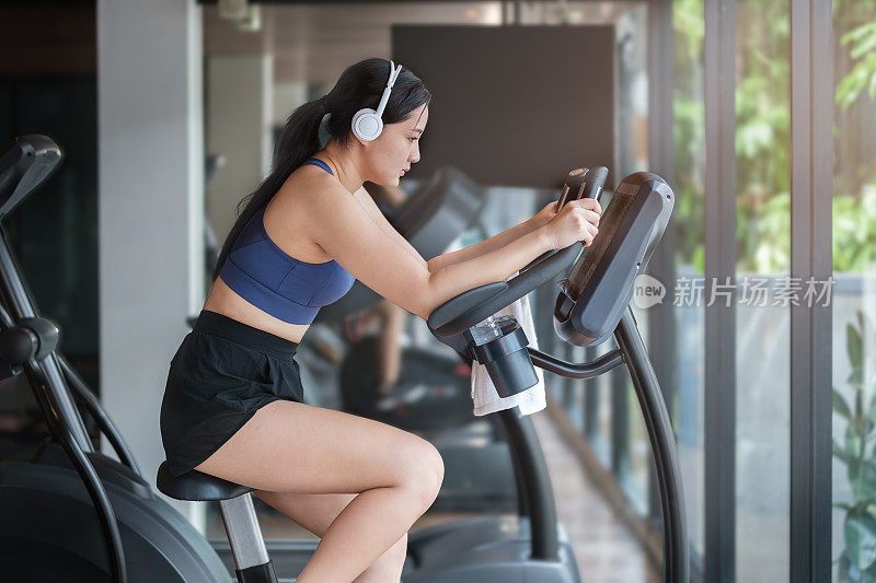 穿着运动服的年轻女子戴着耳机听着音乐。在健身房骑动感单车燃烧卡路里。