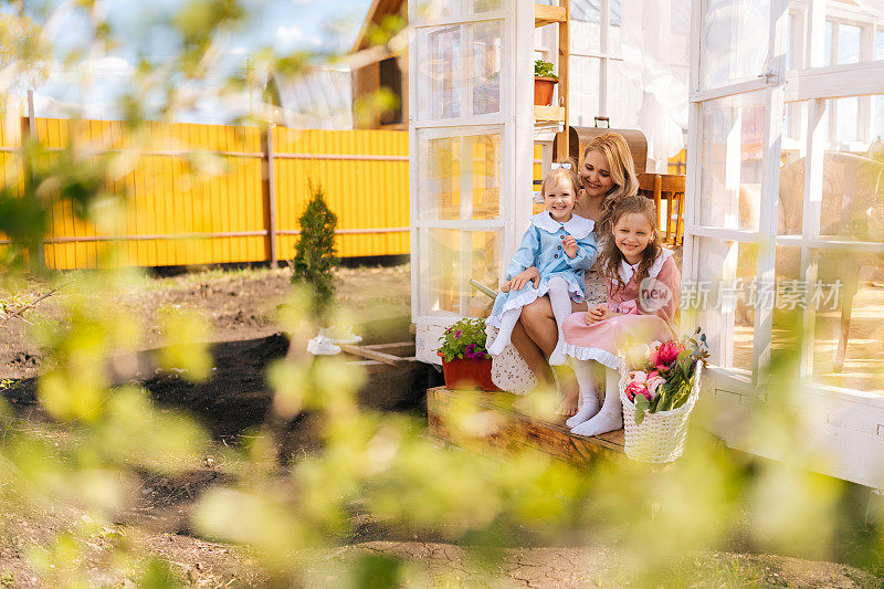 在阳光明媚的日子里，开朗的金发碧眼的年轻母亲和两个可爱的小女儿坐在夏日凉亭的台阶上