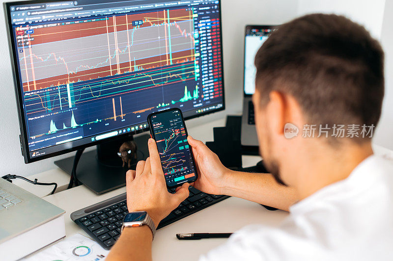 加密货币交易员坐在他的工作场所前的显示器与图表。男性手中的智能手机，屏幕上显示交易数据指数图表，手机应用程序分析加密货币市场