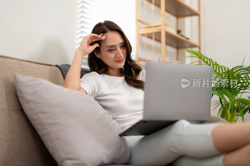 一位年轻女子懒洋洋地躺在家里的沙发上，舒舒服服地使用着她的笔记本电脑，享受着悠闲的一天。