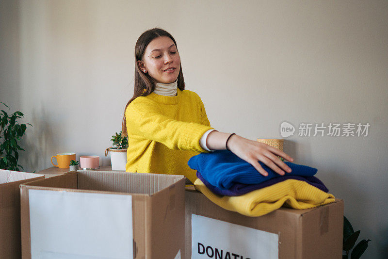 穿着黄色毛衣的年轻女子拿着衣服，在她的房间里放着捐款箱，表达着捐赠的概念