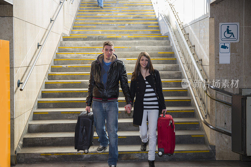 一对年轻夫妇提着行李箱从楼梯走到铁路地下通道