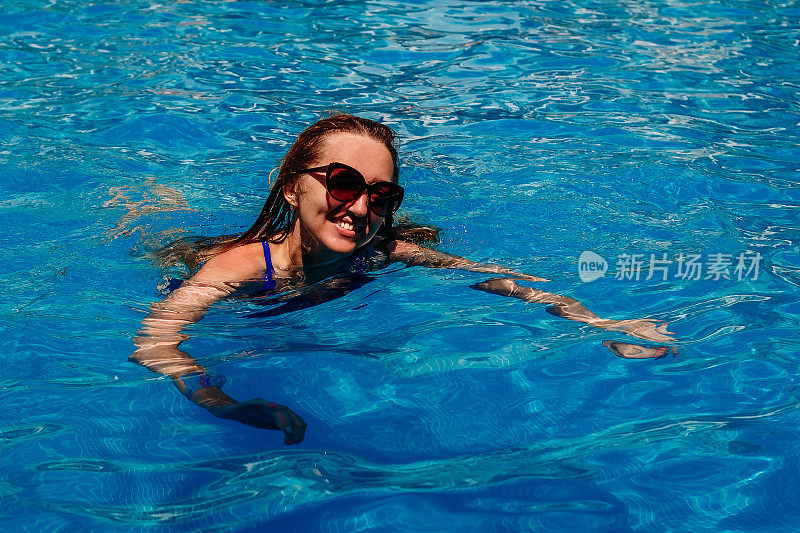 一位穿着泳衣、戴着墨镜、面带微笑的年轻女子在室外游泳池的蓝色水中游泳。