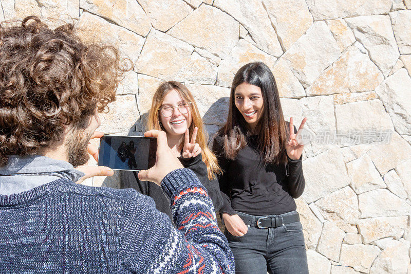 两个年轻女孩在朋友的智能手机镜头前摆姿势，手指排成胜利的V字形——朋友之间的回忆