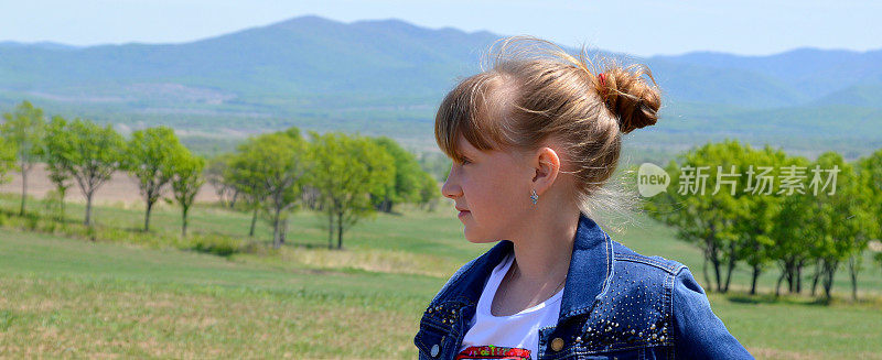 肖像10岁的俄罗斯金发女孩微笑的发髻在绿色的田野与树木的背景。可爱的脸，向左看