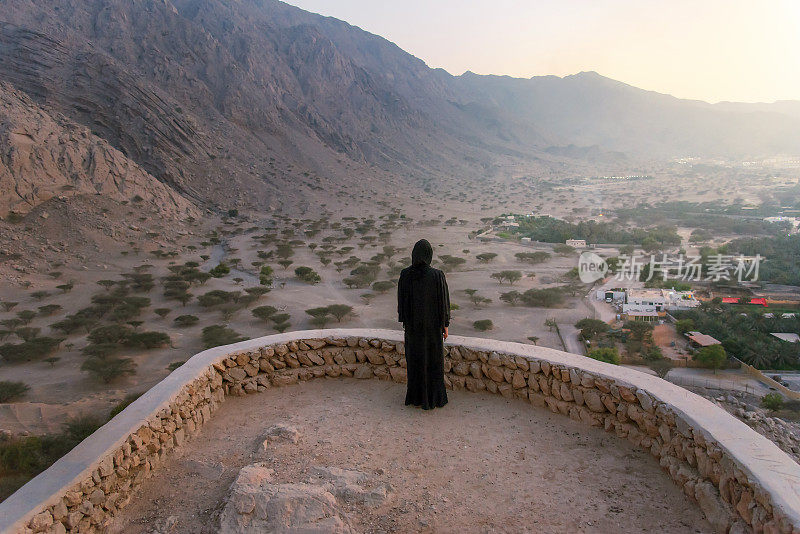 日落时分，一名戴着头巾的妇女在阿联酋北部的沙漠中参观中东的达亚堡