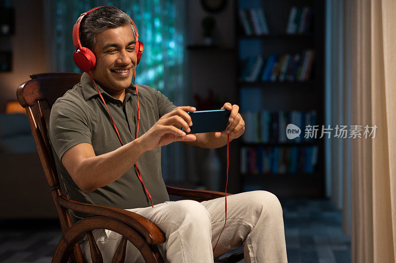 一个快乐的成熟男人坐在摇椅上听音乐，用耳机在家:-股票照片