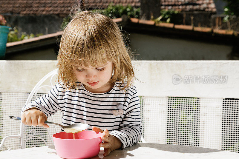 可爱的小孩坐在餐桌旁，在阳台上用粉红色的碗吃饭，微笑着吃早餐