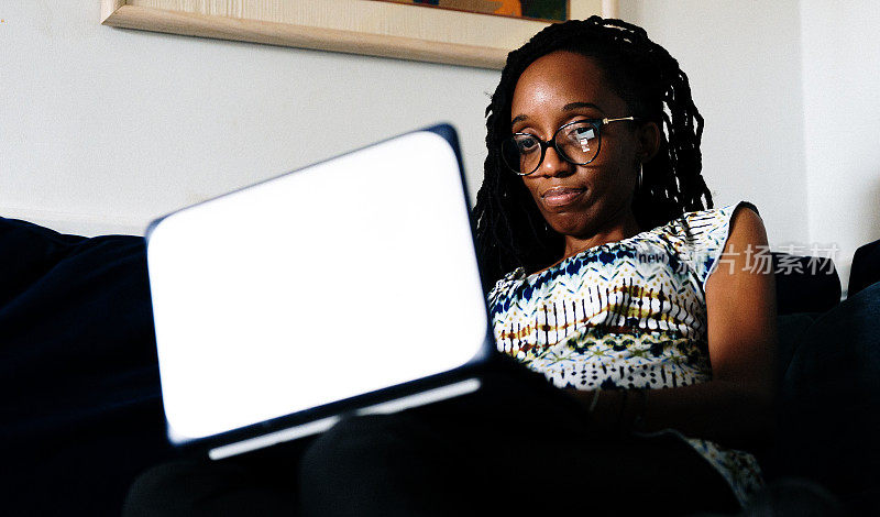 一个开朗的黑人妇女，在家里的沙发上用笔记本电脑放松，她是平静而满足的