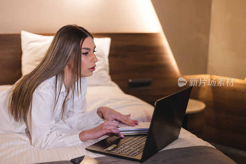 年轻的女商人躺在酒店房间里，一边用笔记本电脑工作，一边在笔记本上记笔记