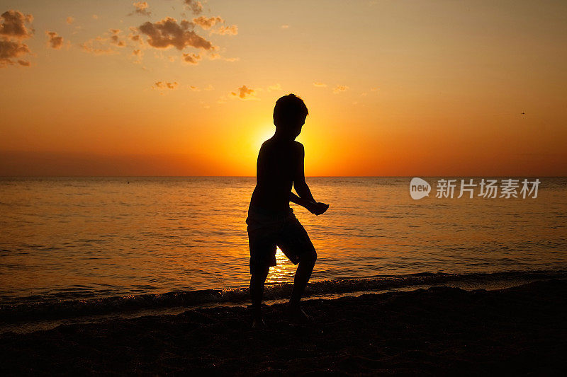 一个夏天的傍晚，一个小男孩走在大海和日落的背景下，手里拿着什么东西在沙滩上