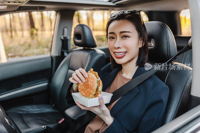 亚洲女人在车里休息，吃汉堡。一个年轻成功快乐的女人正在开车。