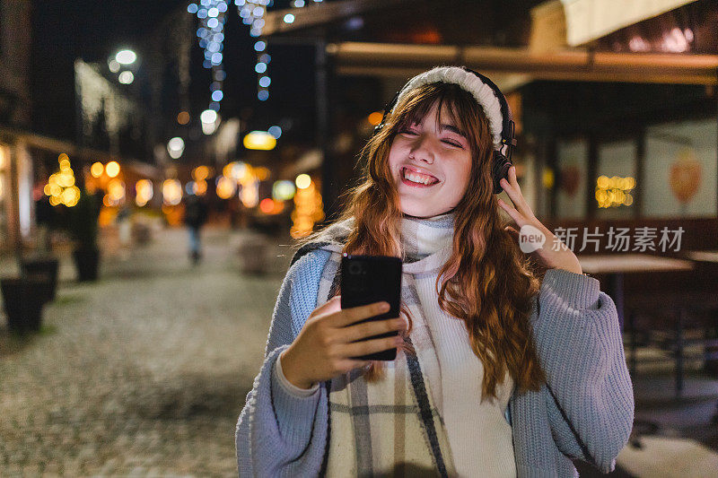 在城市的夜晚，微笑的年轻女子用她的智能手机听音乐