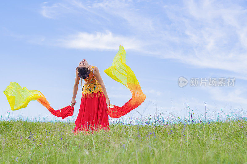 身着红色民族服饰的美女，在蓝天下的绿色田野上跳着东方舞蹈