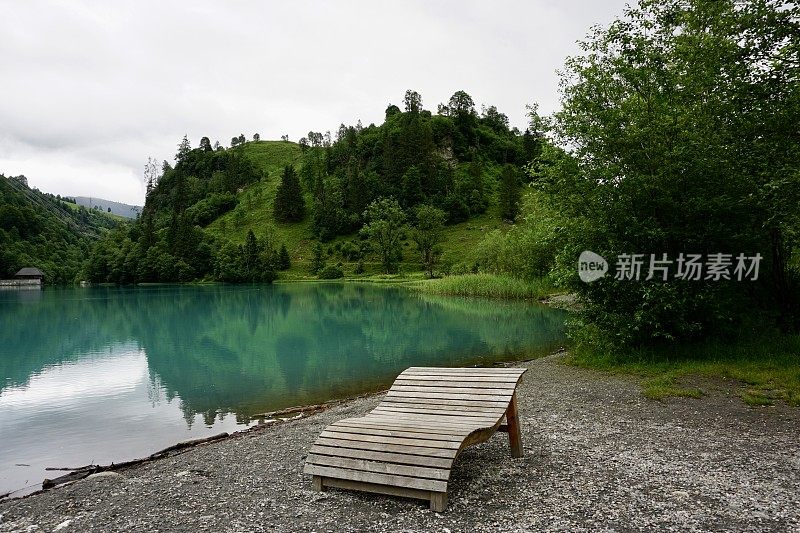 绿松石湖和群山岸边的长凳。在阴天，山附近的绿松石湖。湖边的双木质躺椅。奥地利的克拉姆西湖。明亮的蓝绿色湖泊。