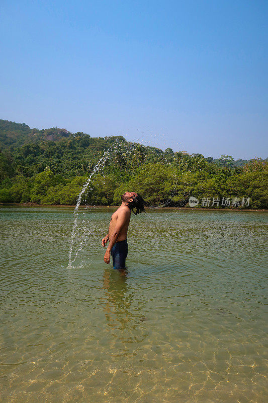 穿着冲浪短裤的印度男子站在齐膝深的泻湖平静的水中，拨弄湿的头发溅起水花，热带岛屿天堂，看到有棕榈树的林地，侧影，聚焦于前景