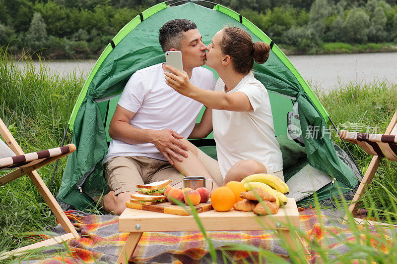 大自然中的快乐时光。在河边露营。浪漫的男女穿着休闲的衣服坐在河边的帐篷里，接吻和自拍。