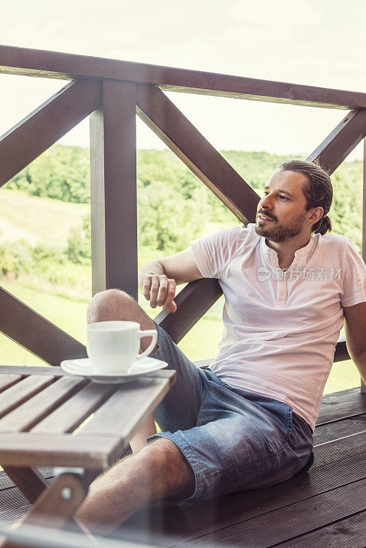 男人坐在阳台的木地板上，喝着咖啡放松