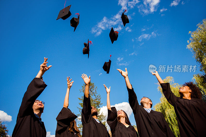 恭喜你!低角度的快乐组的六个年轻的兴高采烈的多民族毕业生在黑色长袍的空中抛起他们的帽子，庆祝，欢笑，享受