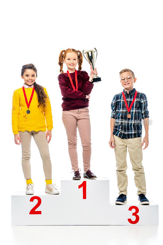 微笑的十多岁的孩子与奖章和奖杯站在获胜者基座上，微笑着看相机孤立在白色