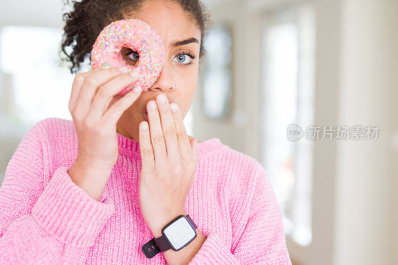 年轻的非洲裔美国女孩吃着甜的粉色甜甜圈，用手捂着嘴，因错误而感到震惊，表情恐惧，在沉默中害怕，秘密的概念