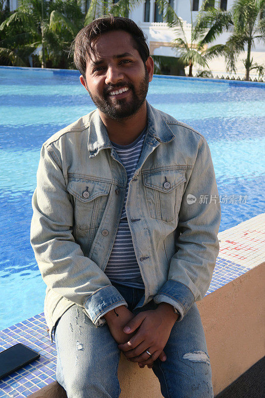 英俊的年轻印度男子坐在游泳池在阳光下看起来快乐的暑假，牙齿微笑，大笑，手在头发，好看的印度男子享受阳光无限游泳池蓝色马赛克瓷砖，度假模式在牛仔夹克