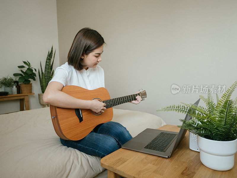 年轻女子微笑着坐在床上拿着吉他看着笔记本电脑